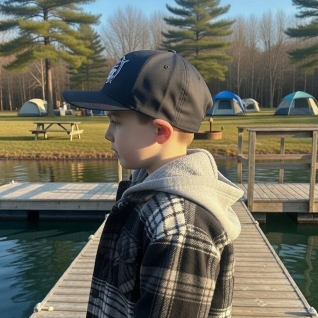 A young boy in a plaid jacket and an AMD Sport-Tek Dry Zone Nylon Cap by American Muscle Docks stands on a dock by a lake, with tents and pine trees in the background on a clear day.