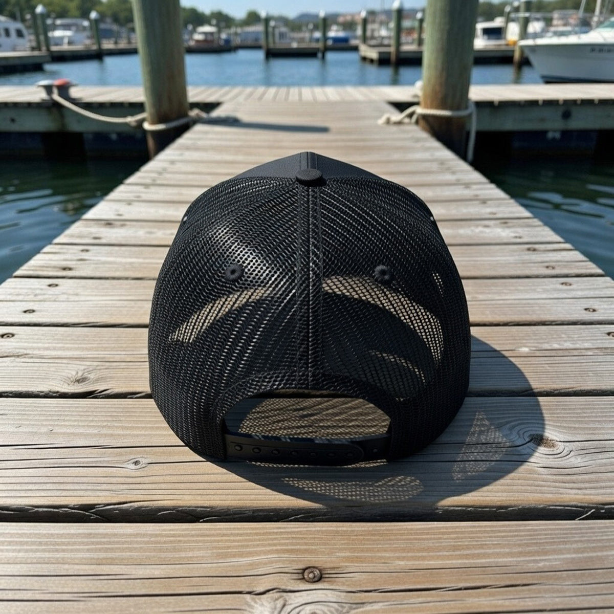 Black mesh American Muscle Docks cap on a wooden dock with water and boats in the background
