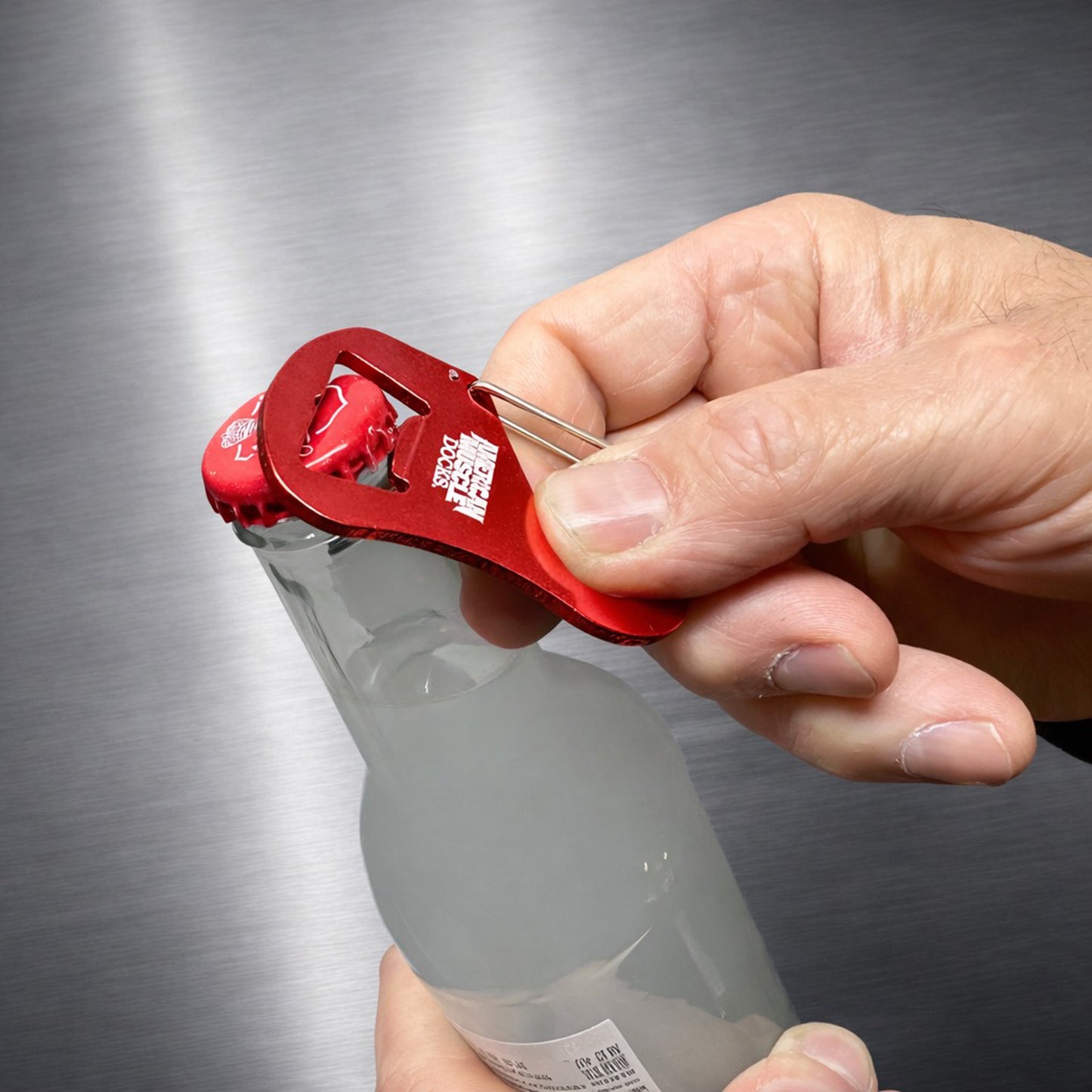 A hand uses the American Muscle Docks Carabiner Bottle Cap Opener to easily remove the cap from a glass bottle, highlighting the convenience of this versatile tool.