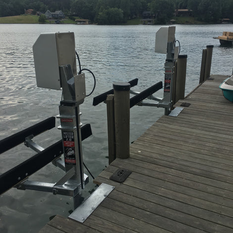 Two metal boat lift mechanisms are installed on a wooden dock by a lake, with trees and houses visible on the opposite shore under a partly cloudy sky.