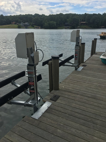 Two metal boat lift mechanisms are installed on a wooden dock by a lake, with trees and houses visible on the opposite shore under a partly cloudy sky.