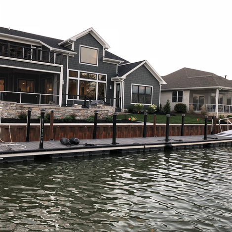 Two modern lakeside houses with large windows and porches are shown, with a long floating dock extending from the shore over the water in front of the homes.