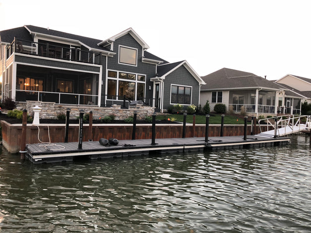 Two modern lakeside houses with large windows and porches are shown, with a long floating dock extending from the shore over the water in front of the homes.