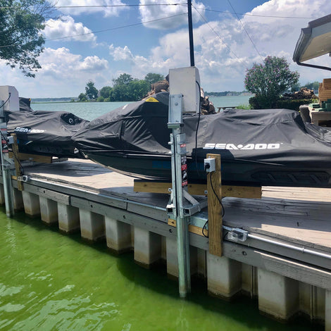 Two covered Sea-Doo jet skis rest on an American Muscle Docks & Fabrication Mr. Lifter - Electric 9' Pipe Track lift by a dock over green water near a house, with trees and a cloudy sky in the background.