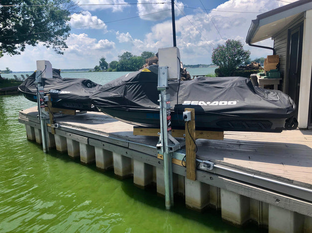 Two covered Sea-Doo jet skis rest on an American Muscle Docks & Fabrication Mr. Lifter - Electric 9' Pipe Track lift by a dock over green water near a house, with trees and a cloudy sky in the background.