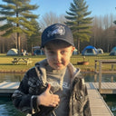 A young boy in a plaid jacket and an AMD Sport-Tek Dry Zone Nylon Cap by American Muscle Docks stands on a dock, giving a thumbs up, with tents and trees in the background.
