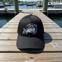 The AMD Sport-Tek Dry Zone Nylon Cap by American Muscle Docks is a black, sweat-resistant cap featuring a stylized swordfish logo, shown on a wooden dock with water and boats in the background.