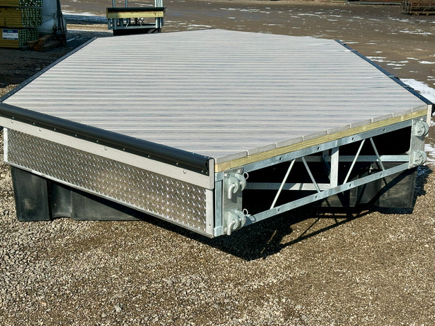 A metal and wood dock platform with a hexagonal shape is positioned on gravel outdoors, featuring metal support beams and a diamond plate side panel.