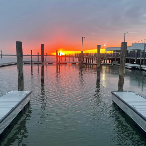 Empty boat docks and wooden piers over calm water at sunset, with an orange sun low on the horizon and a partly cloudy sky.