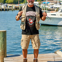 A man stands on a dock with fishing rods, wearing an American Muscle Docks AMD Eagle Crest T-Shirt, fishing vest, cargo shorts, boots, and a cap. Boats and water are seen in the background.