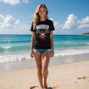 A woman in an AMD Eagle Crest T-Shirt from American Muscle Docks and denim shorts stands on a sandy beach with waves and a partly cloudy sky, embodying American-built pride.