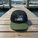 An American Muscle Docks New Era® Camo Flat Bill Snapback - Green, featuring a camo pattern, sits on a wooden dock with water and boats in the background.