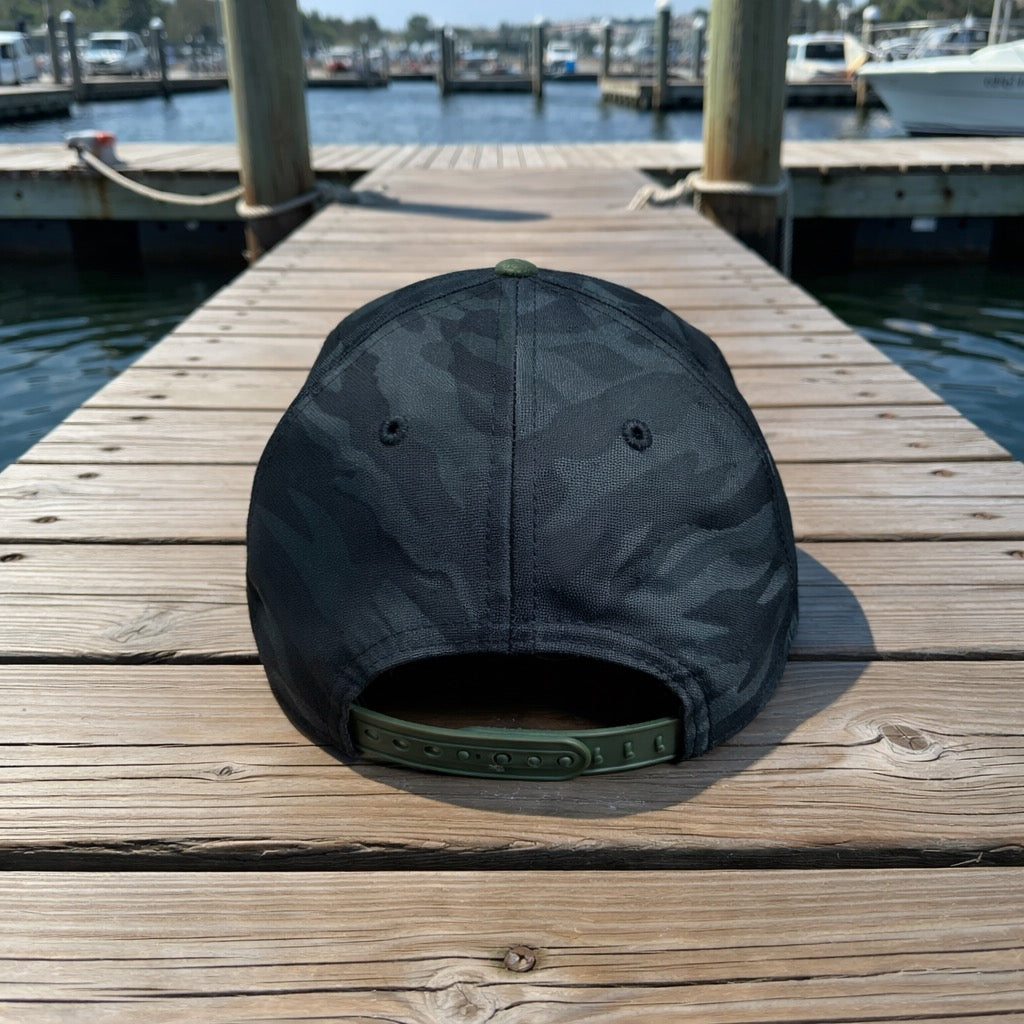 The New Era® Camo Flat Bill Snapback - Green from American Muscle Docks is displayed on a wooden dock with boats and water in the background.