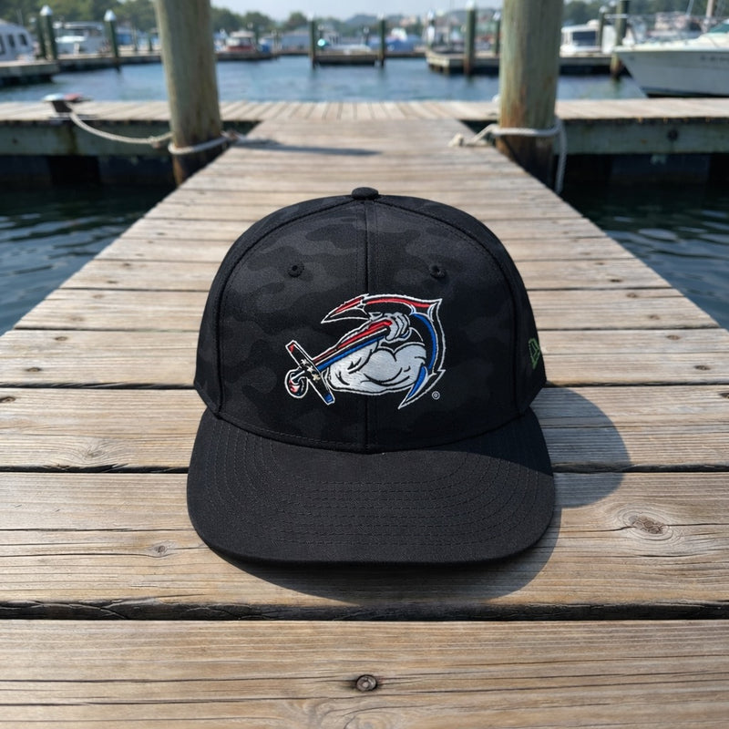 A green New Era® Camo Flat Bill Snapback by American Muscle Docks sits on a wooden dock near the water, with boats and pilings in the background.