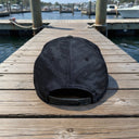 The New Era® Camo Flat Bill Snapback - Green from American Muscle Docks sits upside down on a wooden dock with boats and water behind it, capturing the classic atmosphere of the brand.