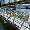 A metal gangway extends over river water, leading to a floating dock, with a yellow bridge and city buildings visible in the background.