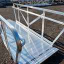 A metal accessibility ramp with handrails on both sides is positioned on a gravel surface outdoors, leading to a floating dock.