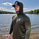 A man wearing the American Muscle Docks AMD Cooling Performance UPF Tee (L/S) with Hood and a cap stands by a lake, surrounded by trees under a blue sky.