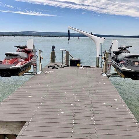 Two jet skis are suspended above the water by mechanical lifts on either side of a wooden dock extending out into a lake.