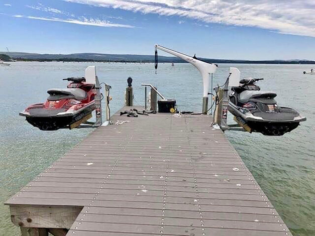 Two jet skis are suspended above the water by mechanical lifts on either side of a wooden dock extending out into a lake.