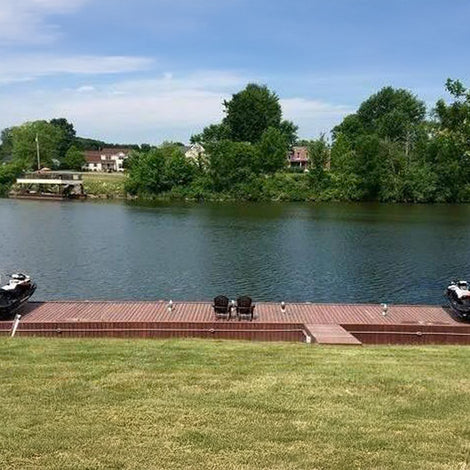 A wooden dock with two fishing boats is on a calm river, with grassy banks in the foreground and trees and houses across the water.