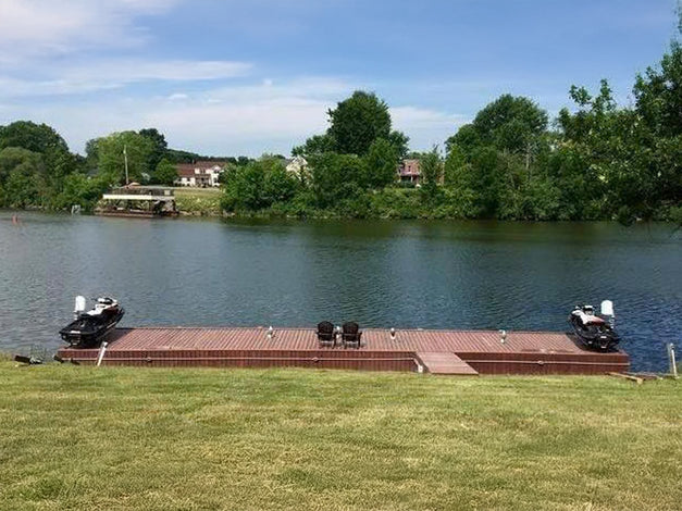 A wooden dock with two fishing boats is on a calm river, with grassy banks in the foreground and trees and houses across the water.