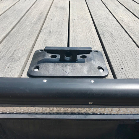 Close-up of a black dock cleat mounted on a wooden dock surface, with part of a black edge bumper and water visible at the bottom.
