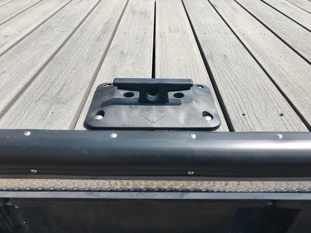 Close-up of a black dock cleat mounted on a wooden dock surface, with part of a black edge bumper and water visible at the bottom.