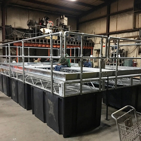 Large metal platform structure with railings sits atop black plastic tanks inside an industrial workshop; workers are present in the background.