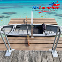 A kayak sits on a wooden dock with a metal launch device, paddle inside, and turquoise water behind. Stable dock entry is ensured by the Mr. Launcher Kayak Launch Dock from American Muscle Docks & Fabrication, logo at top right.