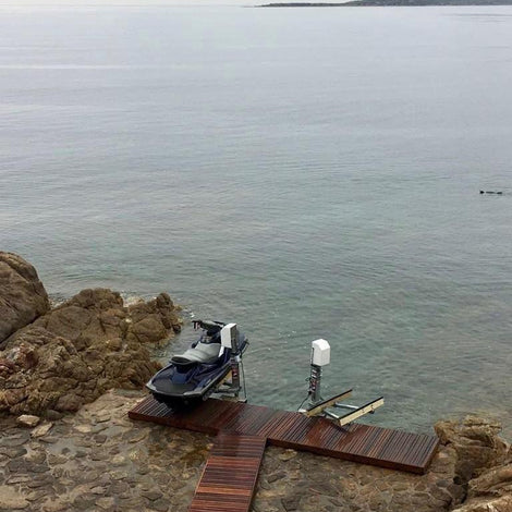 A jet ski is parked on a small wooden dock by rocky shore with calm water and a distant mountain under a cloudy sky.