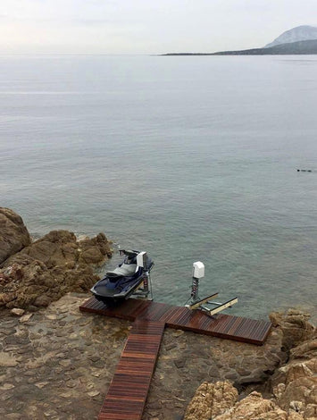 A jet ski is parked on a small wooden dock by rocky shore with calm water and a distant mountain under a cloudy sky.