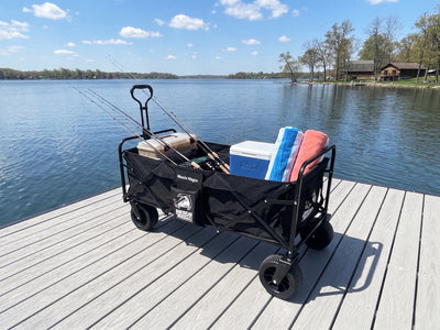 The American Muscle Docks Muscle Wagon, in black, sits on a dock holding fishing rods, a tackle box, a cooler, and towels with a scenic lake and trees in the background beneath partly cloudy skies.