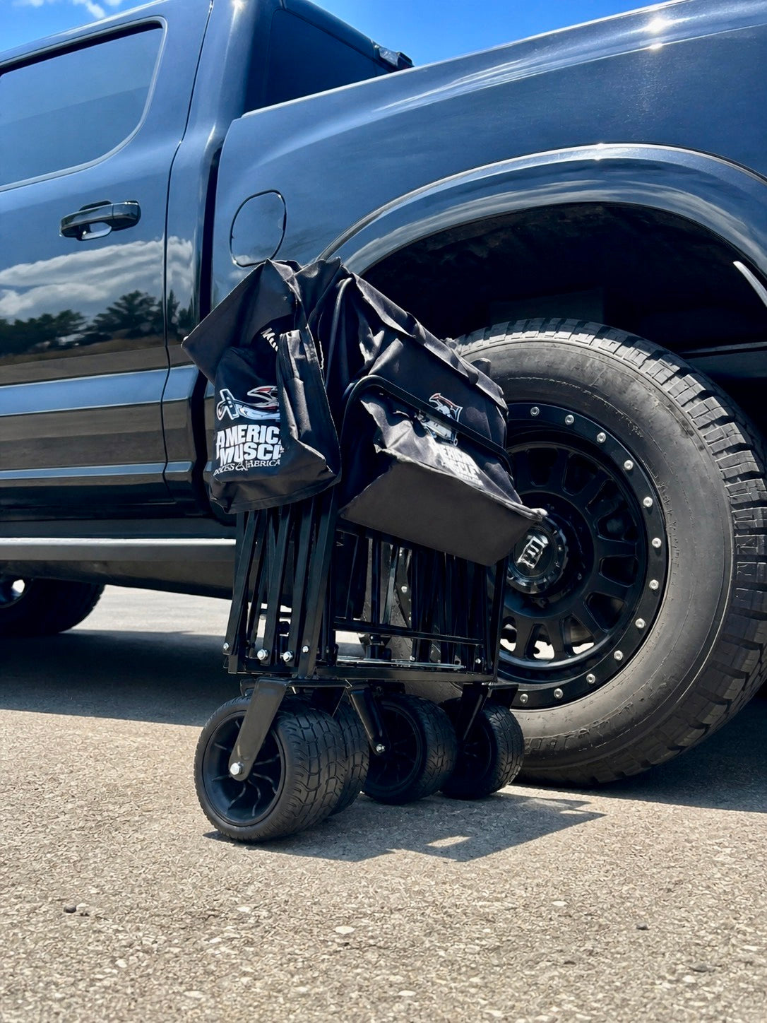 An American Muscle Docks Collapsible Pull Wagon stands folded upright on the pavement next to the rear wheel of a black pickup truck.