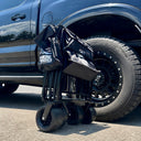 An American Muscle Docks Collapsible Pull Wagon stands folded upright on the pavement next to the rear wheel of a black pickup truck.