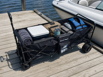 The American Muscle Docks Collapsible Pull Wagon sits on a wooden dock, loaded with towels, a cooler, bags, two life jackets, and a tumbler beside a docked boat.