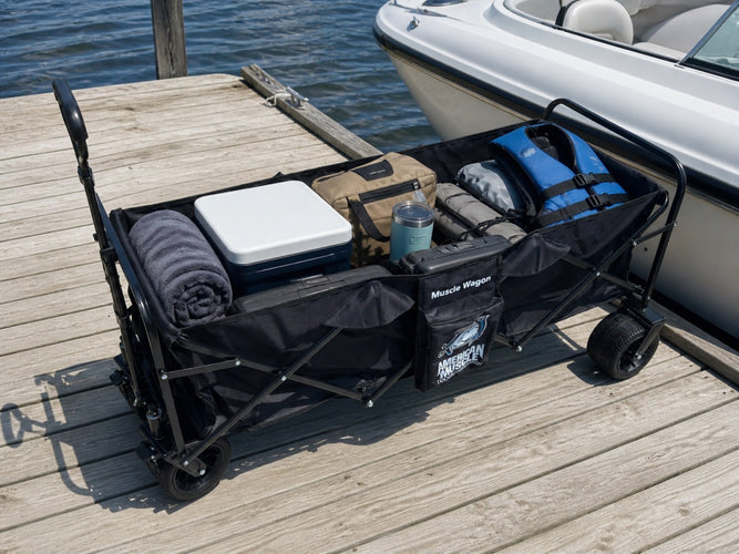 The American Muscle Docks Collapsible Pull Wagon sits on a wooden dock, loaded with towels, a cooler, bags, two life jackets, and a tumbler beside a docked boat.