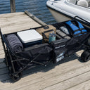 The American Muscle Docks Collapsible Pull Wagon sits on a wooden dock, loaded with towels, a cooler, bags, two life jackets, and a tumbler beside a docked boat.