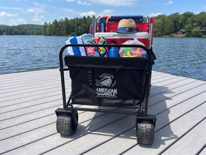 An American Muscle Docks Collapsible Pull Wagon, filled with towels, a hat, water bottles, and chairs, rests on a dock by a lake with trees and cabins in the background.
