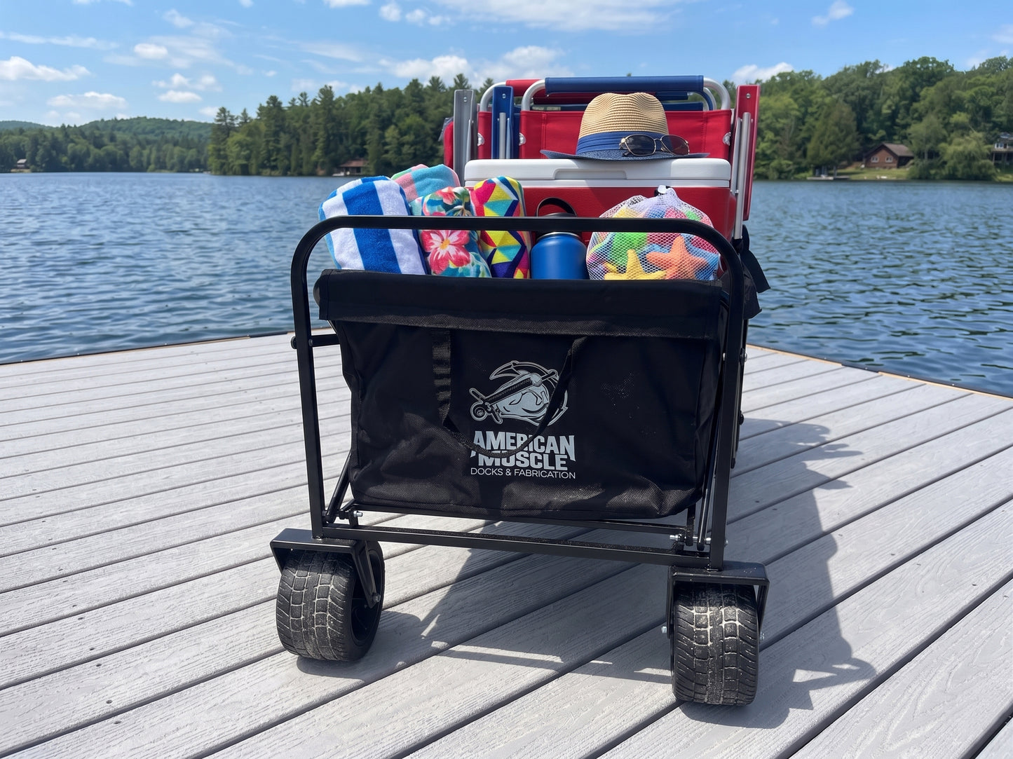 An American Muscle Docks Collapsible Pull Wagon, filled with towels, a hat, water bottles, and chairs, rests on a dock by a lake with trees and cabins in the background.
