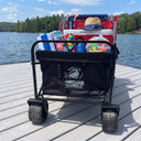 An American Muscle Docks Collapsible Pull Wagon, filled with towels, a hat, water bottles, and chairs, rests on a dock by a lake with trees and cabins in the background.