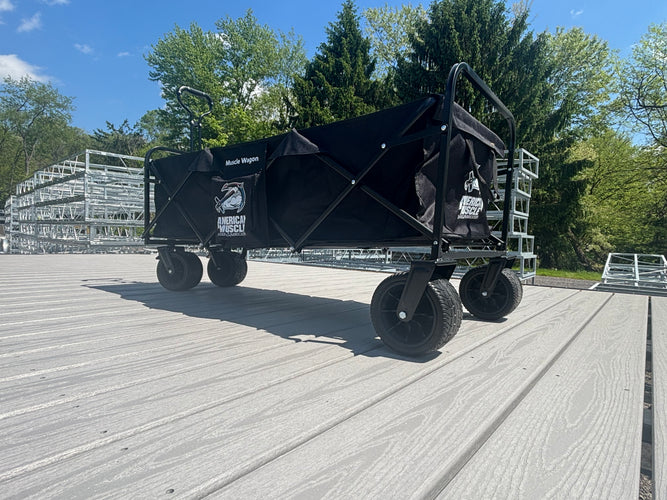 A black Collapsible Pull Wagon by American Muscle Docks stands on a wooden deck outdoors, with trees and steel truss dock frames in the background.