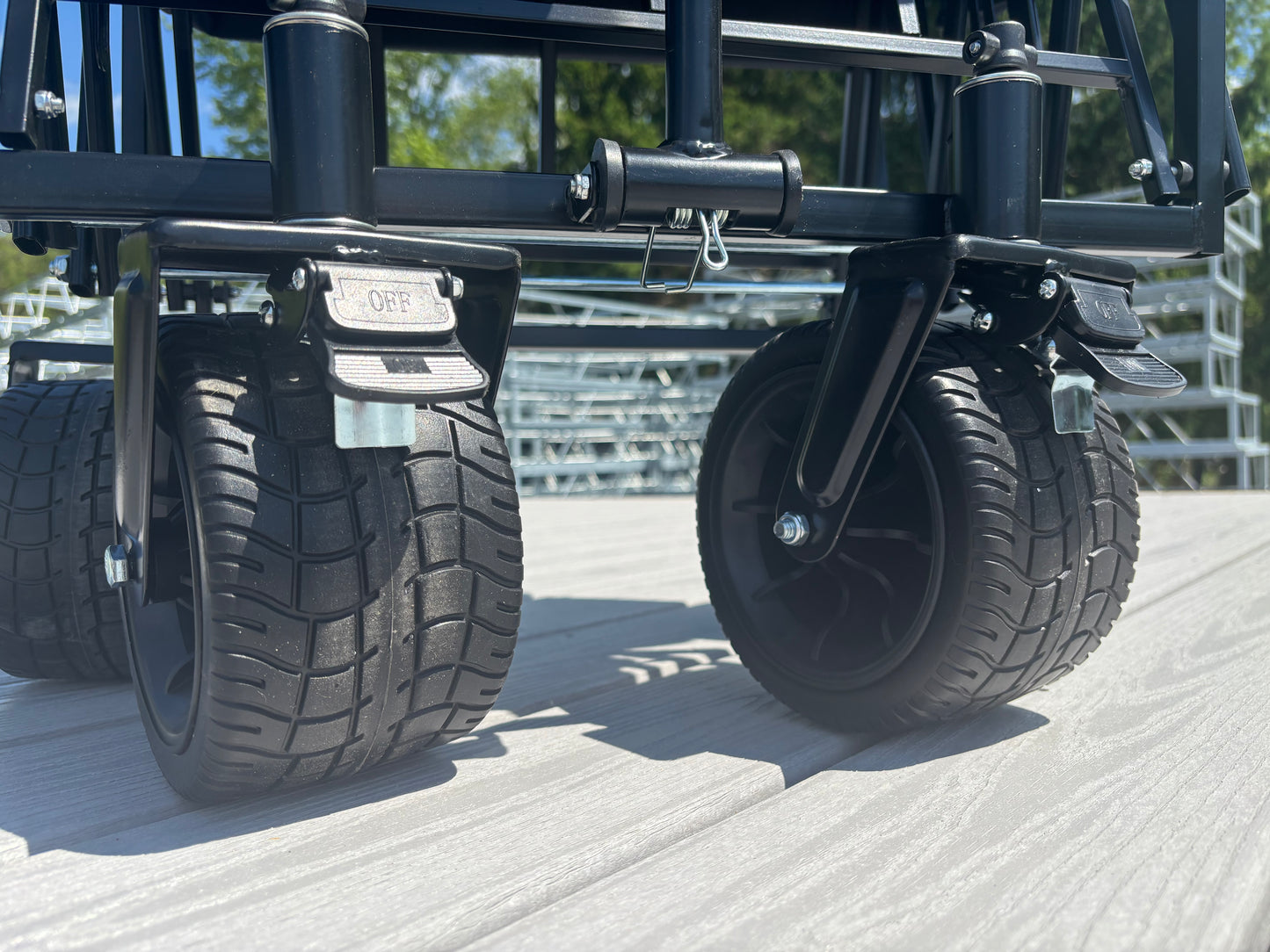 Close-up of two large black treaded caster wheels attached to the metal frame of an American Muscle Docks Collapsible Pull Wagon on a light-colored outdoor deck.