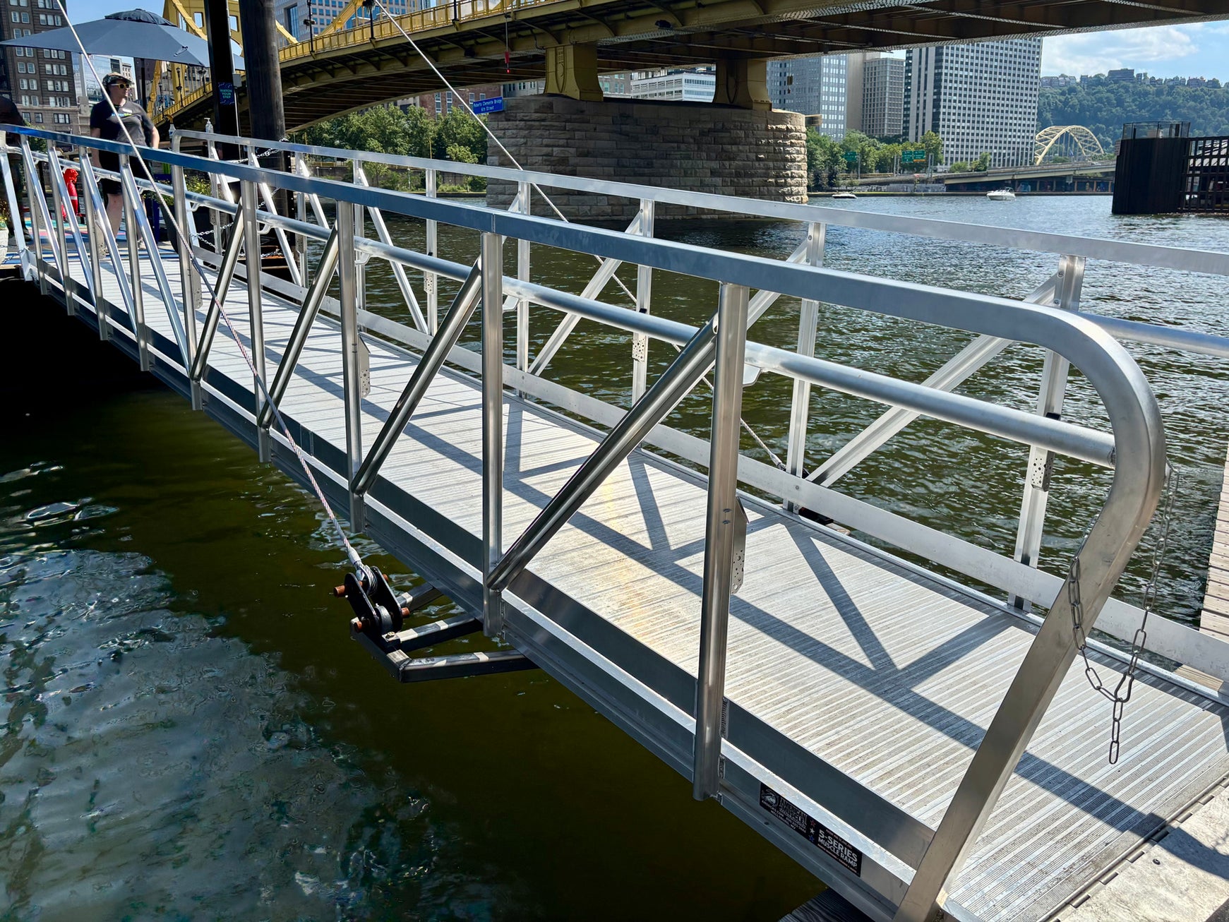 A metal gangway bridge extends over a river, connecting a floating dock to the shore. In the background, a yellow bridge and city buildings are visible.