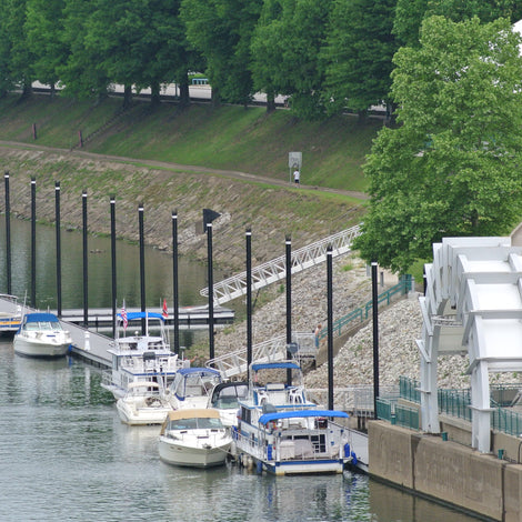 Several boats are docked along a riverside marina, with a paved walkway, green trees, and a white canopy structure nearby.