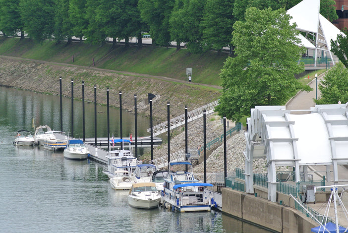 Several boats are docked along a riverside marina, with a paved walkway, green trees, and a white canopy structure nearby.