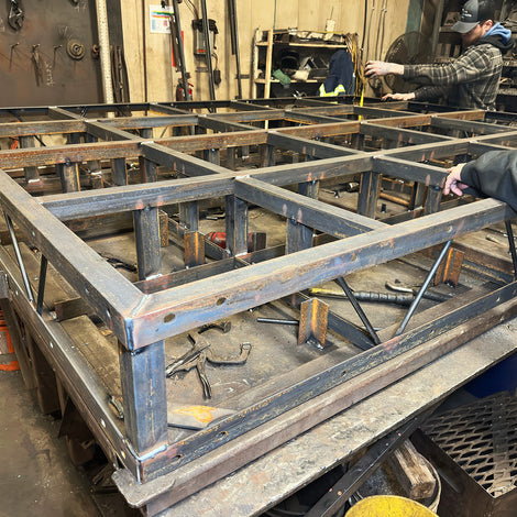 Two people work on a large metal frame structure for a floating dock in a workshop, with metal tools and materials visible in the background.