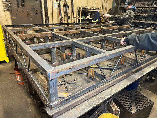 Two people work on a large metal frame structure for a floating dock in a workshop, with metal tools and materials visible in the background.