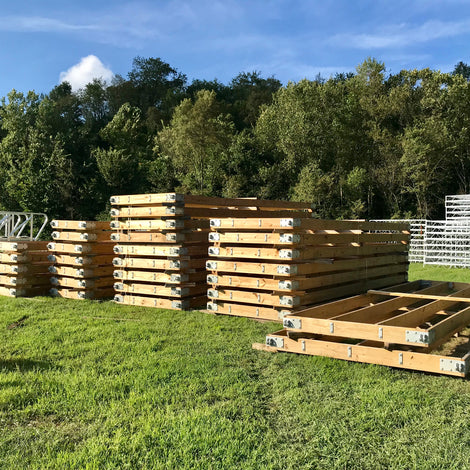 Stacks of wooden beams with metal brackets are arranged on grass near trees under a partly cloudy sky.