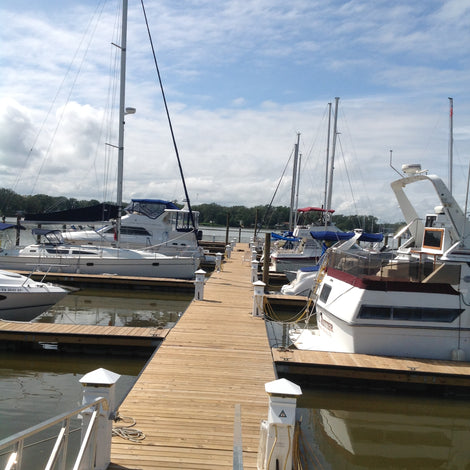 A wooden dock extends into the water with several boats and yachts moored on both sides under a partly cloudy sky.